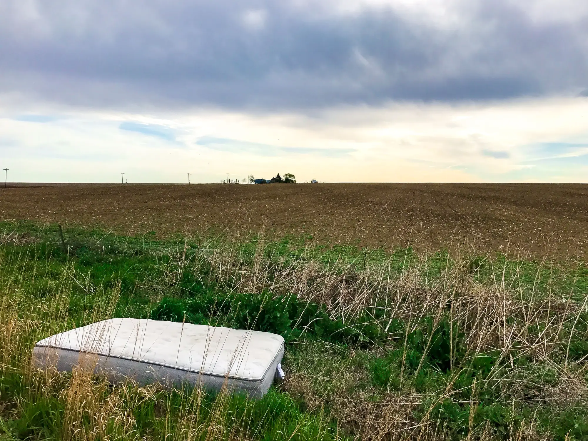 An old mattress dumped in the long grass at the edge of a ploughed field, under a grey sky